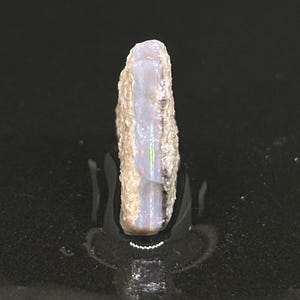 May include: A rough, white, and gray opal with iridescent flashes of color. The opal is a long, thin, and rectangular shape.