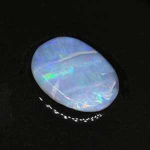 May include: A polished, oval-shaped opal gemstone. The opal displays a milky white base color with iridescent flashes of blue, green, and yellow. The gemstone is set against a black background, highlighting its vibrant play-of-color.