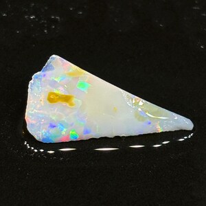 Könnte beinhalten: Ein weißer Opal-Edelstein mit schillernden Regenbogenfarben und einer kleinen gelben Einlagerung. Der Opal ist ein roher, natürlicher Stein mit einer unregelmäßigen, dreieckigen Form.