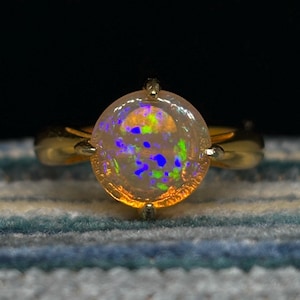 May include: A gold ring with a round opal gemstone. The opal is a light pink color with green, blue, and purple flashes of color.