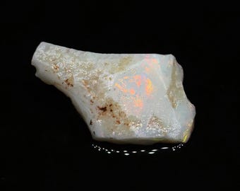 May include: A raw opal gemstone, white in colour with internal flashes of orange and yellow. The opal is irregularly shaped and has a textured surface. The gemstone is displayed against a black background.