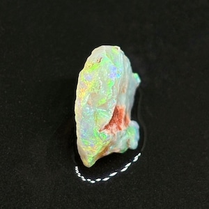 Könnte beinhalten: Ein roher, weißer Opal mit schillernden grünen, blauen und roten Blitzen. Der Opal ist ein natürlicher Edelstein und hat eine raue, unregelmäßige Form.