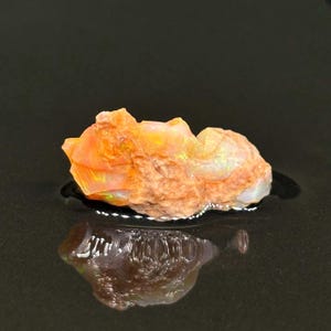 Puede incluir: Una piedra preciosa de ópalo en bruto, de color naranja y blanco, con una forma natural irregular. El ópalo está sobre una superficie negra y refleja la luz.
