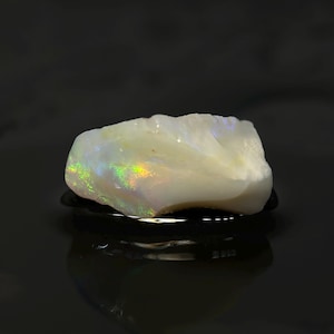 May include: A raw, uncut opal gemstone. The opal is primarily white with iridescent flashes of green, yellow, and blue. The stone is set against a dark, reflective surface, highlighting its natural beauty and unique color play.