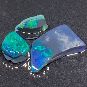 May include: Three pieces of raw opal, one is a dark blue with green flashes, one is a light blue with green flashes, and one is a dark green with blue flashes.