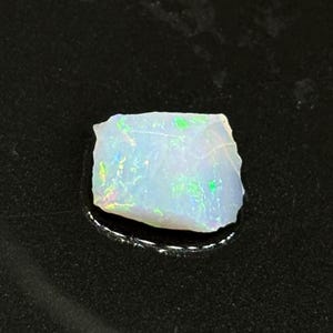 Op de afbeelding: Een witte opaal edelsteen met groene en blauwe irisatie. De opaal is ruw en heeft een onregelmatige vorm.