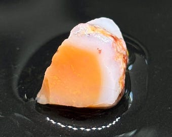 May include: A rough, orange and white opal gemstone with a translucent, milky white centre and a reddish-orange outer layer. The stone is sitting on a black surface.