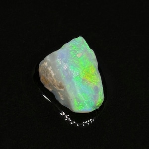 May include: A raw opal gemstone with a rough, irregular shape. The stone displays a mix of colors, including white, green, and yellow, with a shimmering, iridescent quality. The opal is set against a dark background.