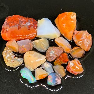 Könnte beinhalten: Eine Sammlung von rohen Opal-Steinen in verschiedenen Schattierungen von Orange, Weiß und Grau. Die Steine sind auf einer schwarzen Oberfläche angeordnet.