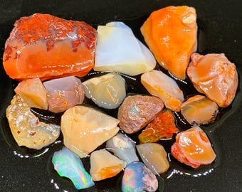 May include: A collection of rough opal stones in various shades of orange, white, and grey. The stones are arranged on a black surface.
