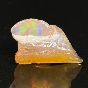 Puede incluir: Un pequeño ópalo áspero, blanco y naranja con un brillo iridiscente arcoíris.