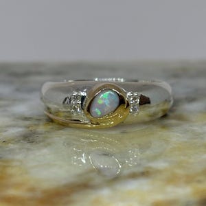 May include: A silver and gold ring featuring an oval opal gemstone with iridescent colors. The ring has a gold accent and small diamond-like stones on either side of the opal. The ring is a piece of jewelry.