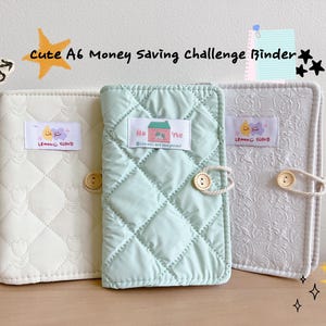 May include: Three A6 money-saving challenge binders in cream, mint green, and white. The cream and mint green binders have a quilted pattern, while the white binder has a floral pattern. Each binder has a button closure and a label with a logo.