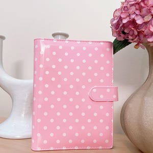 May include: A rectangular pink planner with a white polka dot pattern. The planner features a glossy finish and a matching pink strap closure with a white polka dot design on the right side to secure the cover shut when not in use.