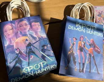 May include: Blue paper gift bags featuring illustrations of anime-style characters. The bags have white paper handles. The text "KPOP DEMON HUNTERS" is printed on the front of the bags. The bags are stacked on a wooden surface.