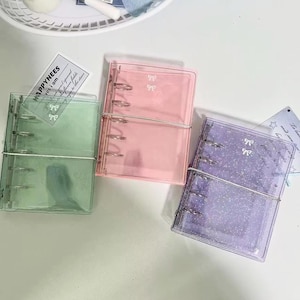 May include: Three clear, colorful binders in green, pink, and purple. Each binder has a silver metal ring binder and bow accents. The binders are on a white surface with small decorative cards.