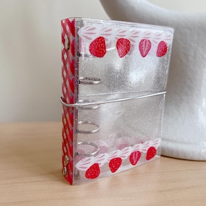 May include: A clear, glittery mini binder with a strawberry design. The binder has a red and white gingham spine and a decorative strawberry pattern along the top and bottom edges. The binder is held together with silver metal rings and a wire closure.