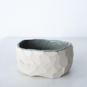 May include: A small, hand-formed ceramic bowl with a matte off-white exterior featuring a faceted, geometric texture. The interior of the bowl is finished in a smooth, muted sage green glaze that extends to the uneven, organic rim of the vessel.