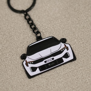 May include: White car-shaped keychain with black accents, including a black keyring and chain. The car has orange accent lights and the letters "KOH" on the front. The keychain is on a neutral-colored surface.