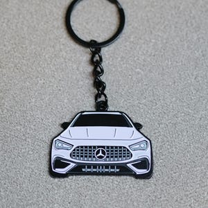 Peut inclure: Porte-clés en forme de voiture blanche avec des bordures noires, une chaîne noire et un anneau noir. L'avant de la voiture présente une calandre et des phares détaillés. Le porte-clés est probablement en métal.