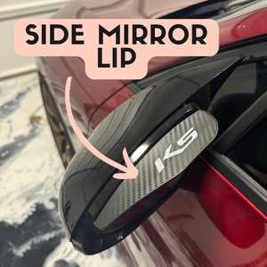 Puede incluir: Primer plano de un espejo retrovisor negro con un labio de espejo retrovisor de aspecto de fibra de carbono. El labio tiene las letras "KS" impresas en blanco.