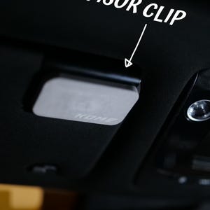 Peut inclure: Un clip de pare-soleil de voiture noir avec le texte "VISOR CLIP" en blanc. Le clip a une plaque rectangulaire de couleur argentée avec le nom de la marque "KOME" imprimé dessus. Le clip est fixé à l'intérieur de la voiture.