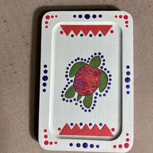 May include: A white wooden tray with a red and blue border. The tray features a green and red turtle design in the center.