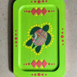 May include: A green wooden tray with a painted design of a sea turtle with a colorful patterned shell. The tray is decorated with red and yellow dots around the edge and in the center.