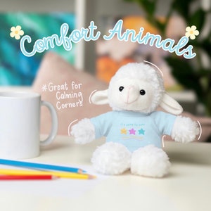 May include: A white plush lamb wearing a light blue shirt with the text "it's okay to feel" and star graphics. The image also includes the text "Comfort Animals" and "Great for Calming Corners". A white mug and colored pencils are in the background.