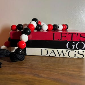 May include: A stack of three decorative wooden books in red, black, and white. The top book is red with the words "LET'S" in black. The middle book is black with "GO" in white. The bottom book is white with "DAWGS" in black. A beaded garland with red, black, and white beads is draped over the books.