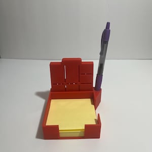 May include: A red desk organizer with a pen holder, a section for small items, and a square section holding yellow sticky notes. A purple and clear pen is in the pen holder. The organizer is on a white surface.