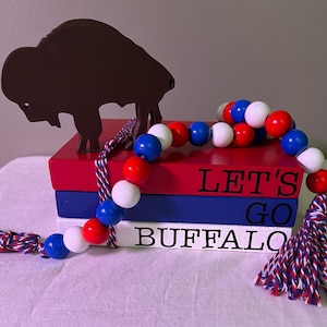 May include: Three wooden blocks stacked on top of each other. The top block is red and says "LET'S GO". The middle block is blue and says "BUFFALO". The bottom block is white and says "BUFFALO". A red, white, and blue beaded garland with a tassel hangs off the side of the blocks. A brown metal buffalo silhouette sits on top of the blocks.