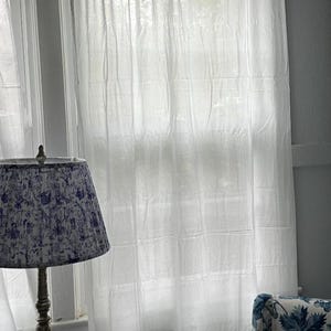 Puede incluir: Escena interior luminosa con cortinas blancas transparentes, una pantalla de lámpara con estampado azul y blanco y una fotografía enmarcada sobre una mesa pequeña. Las cortinas filtran la luz solar.