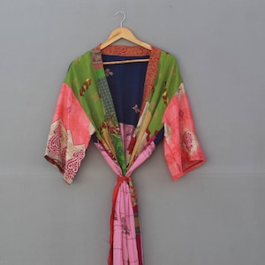 May include: A colorful patchwork kimono robe with a variety of patterns and colors, including pink, green, blue, and red. The robe is hanging on a wooden hanger against a gray background. The robe is tied at the waist with a matching sash.