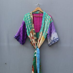 May include: A colorful patchwork kimono robe with a tie closure. The robe features a mix of vibrant colors and patterns, including purple, teal, pink, and floral designs. It is hanging on a wooden hanger against a gray background.
