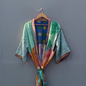 May include: A colorful patchwork kimono robe with a variety of patterns and colors, including blue, green, orange, and pink. The robe is hanging on a wooden hanger against a gray background. The robe is tied at the waist.