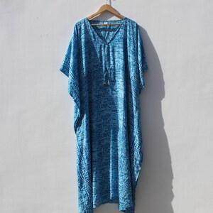 Könnte beinhalten: Ein bodenlanges Kaftan-Kleid mit einem blau-weißen Batikmuster und gitterartigem Motiv. Das Kleidungsstück hat einen V-Ausschnitt, kurze, weite Ärmel und ein Kordelzug-Detail in der Mitte der Brust, präsentiert auf einem Holzkleiderbügel.