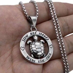 May include: Silver necklace with a round pendant featuring a turtle and the inscription "Slow and steady wins the race".