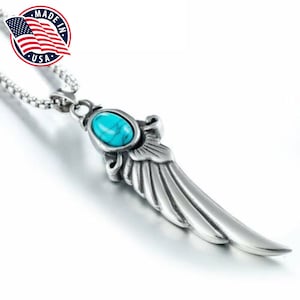Op de afbeelding: Een zilveren ketting met een turquoise steen en een vleugel hanger. De hanger is gedetailleerd met ingewikkelde lijnen en een textuur. De ketting is gemaakt in de VS.