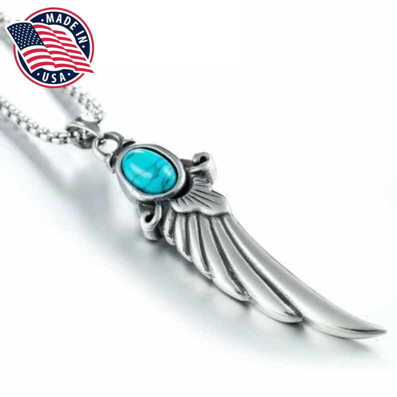 Op de afbeelding: Een zilveren ketting met een turquoise steen en een vleugel hanger. De hanger is gedetailleerd met ingewikkelde lijnen en een textuur. De ketting is gemaakt in de VS.