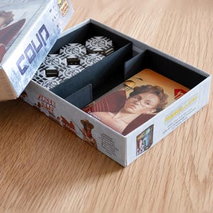 May include: Open game box for the card game "Coup" with a silver and black design. The box contains game cards, black and white game chips, and a card featuring a woman with glasses. The box is on a wooden surface.