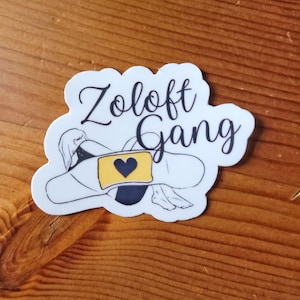 May include: White sticker with the words "Zoloft Gang" in a cursive font. The sticker features a black and white illustration of a person lying down with a bandage over their chest. A yellow rectangle with a blue heart is on the bandage.