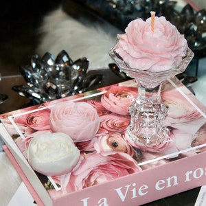May include: Three pink rose-shaped candles, one on a glass pedestal, on a book with the title "La Vie en rose" in pink lettering.