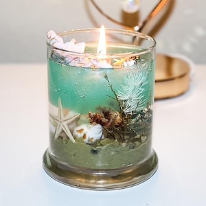 May include: A burning candle in a clear glass jar with a marine theme. The candle has a turquoise wax layer with starfish, shells, and greenery. The jar sits on a gold-coloured base, creating a decorative accent.