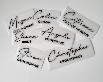 May include: A collection of rectangular, semi-transparent white labels featuring black text. Each label displays a name in a cursive script font above a role in a sans-serif font, including Megan Best Woman, Colin Groom, Catriona Maid of Honour, Shona Bride, Angela Bridesmaid, Steven Groomsman, and Christopher Groomsman.