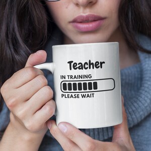 Może przedstawiać: Biały kubek ceramiczny z czarnym tekstem, który brzmi "Teacher in Training" i pasek postępu z napisem "Please Wait".