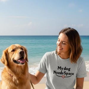 May include: A golden retriever sits on a sandy beach next to a person wearing a gray t-shirt with the text "My dog Needs Space" in black script. The dog is on a leash. The background features the ocean and a blue sky.