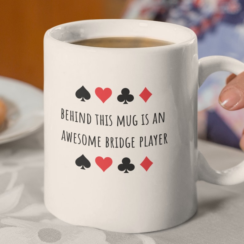 May include: White ceramic coffee mug with black text that reads "Behind this mug is an awesome bridge player". The text is surrounded by four black playing card suits: spades, hearts, clubs, and diamonds.
