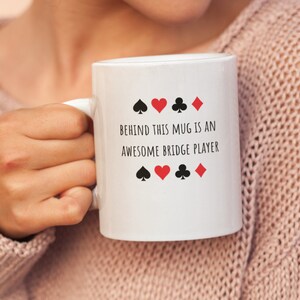 May include: White ceramic mug with black text that reads "Behind this mug is an awesome bridge player". The text is surrounded by four black and red playing card suits.