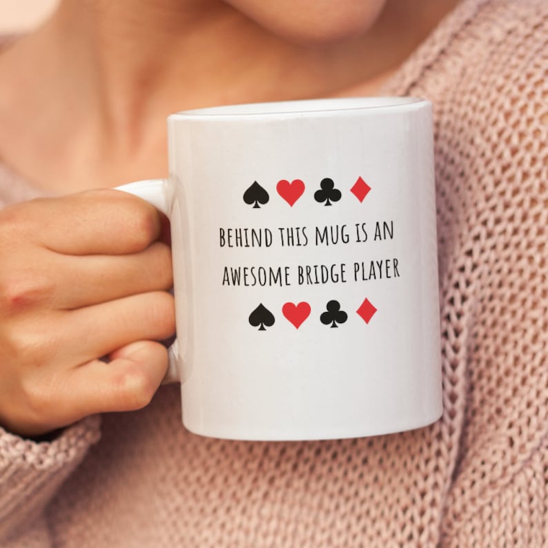May include: White ceramic mug with black text that reads "Behind this mug is an awesome bridge player". The text is surrounded by four black and red playing card suits.